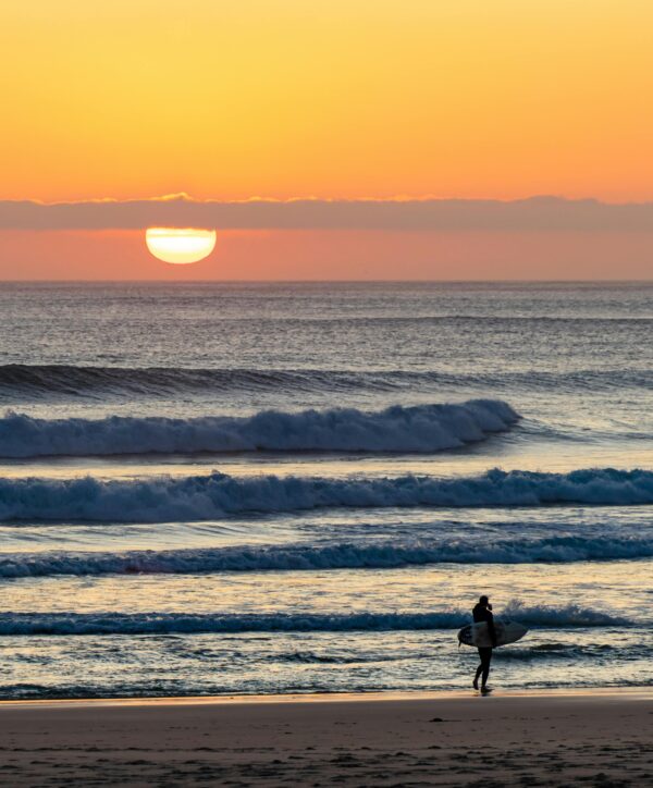 A surfer walks along Costa da Caparica beach at sunset, waves crashing gently on the shore.